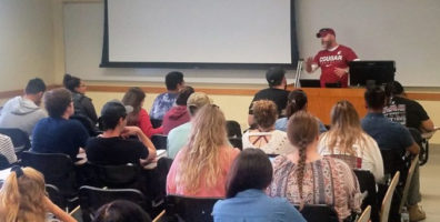 Alumnus Special Agent Craig A. Bowling wears a WSU Cougars t-shirt while talking from the lectern to a group of students in a classroom at WSU Pullman.