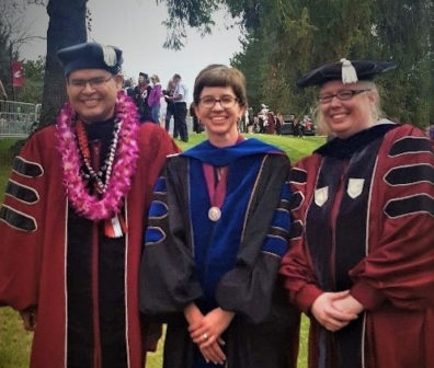 Two graduating PhD students, Jose Collazo, to the left, and Valerie Adrian, to the right, stand with their professor, Julie Kmec, who is standing in the center, all three are wearing commencement regalia and are standing together outside, after the graduation ceremony, all three are smiling at the camera.