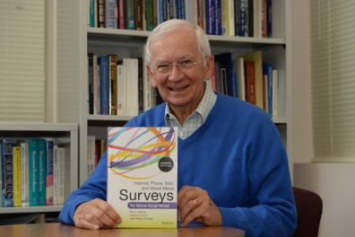 Front facing photo of Don Dillman sitting at a table and holding the text he authored, entitled, "Internet, Phone, Mail, and Mixed Mode Surveys: The Tailored Design Method."
