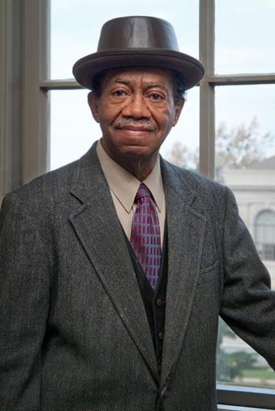 Gordon shown from the waist up, wearing a tweed jacket, white button-down, a tie, and a brown pork-pie hat. He is standing in front of a window, looking into the camera, and smiling.