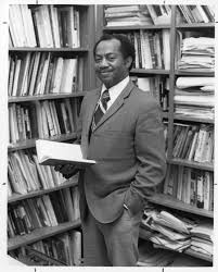 A younger Gordon Morgan shown standing, from the knees up, wearing a suit. Gordon is standing among shelves filled with books and papers. He is holding an open book in one hand, his other hand is in his pocket. He is looking at the camera and smiling. The photo is black and white.