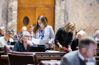 Alex shown standing in the middle of several desks where legislative members are sitting and working. Alex is passing out papers. She is wearing a white jacket and a white, blue, and black blouse.