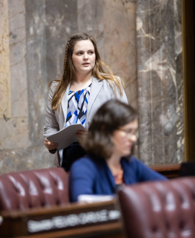 Alex looking away from the camera, walking behind a legislative member, who is sitting. Alex is holding a stack of papers. She is wearing a white jacket, and a white, black, and blue blouse.