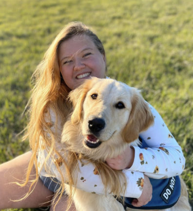 Erika is shown sitting on grass, hugging her golden retriever. Both Erika and her dog are looking at the camera and smiling.
