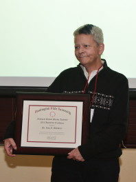 Lisa McIntyre standing, shown from the waist up with a black sweater on. She is holding a frame teaching award given to her at a ceremony.