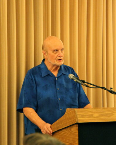 Armand Mauss shown from the waist up speaking at a podium.