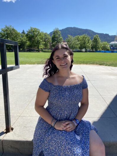 A front facing photo of Georgia Plotkin, from the knees up. She is sitting on cement stairs outside, and is looking into the camera and smiling.