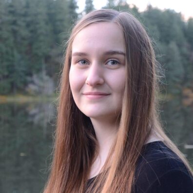 Ally is looking towards the camera and softly smiling, with trees in the background.