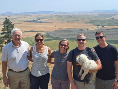 The group is lined up and smiling with their arms around each other. The Palouse is seen behind them.