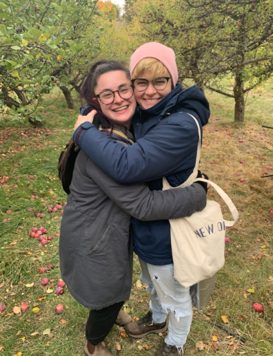 Georgia and Ish are hugging each other and smiling among the apple trees.