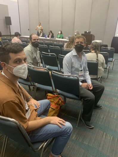 Dylan, Thomas, and Erik are seated in chairs in a large room, masked, and turning towards the camera.