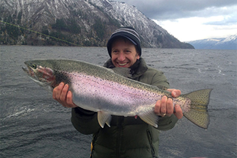 Alex Fremier holding a rainbow trout with Pend Oreille and mountains in the background.
