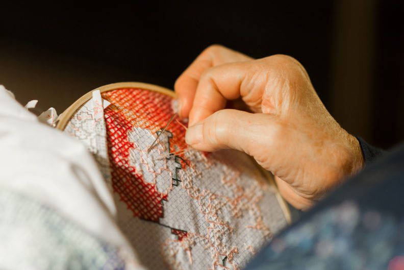 Elderly hands working on cross-stitch needle work.