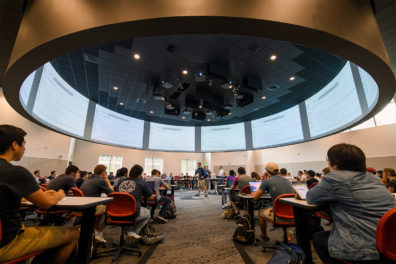 Students sit at desks facing a center projection system while a student speaks in the middle