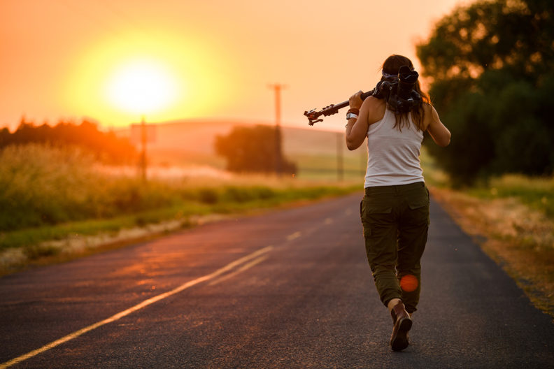 Person walking down road at sunset with camera and tripod over shoulder.