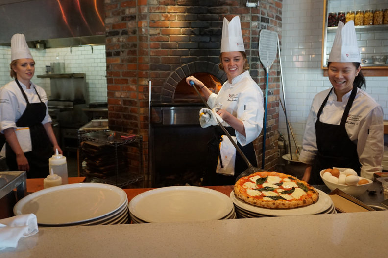 Jacobs takes a pizza out of brick pizza oven as Dahlin and Yuen watch.