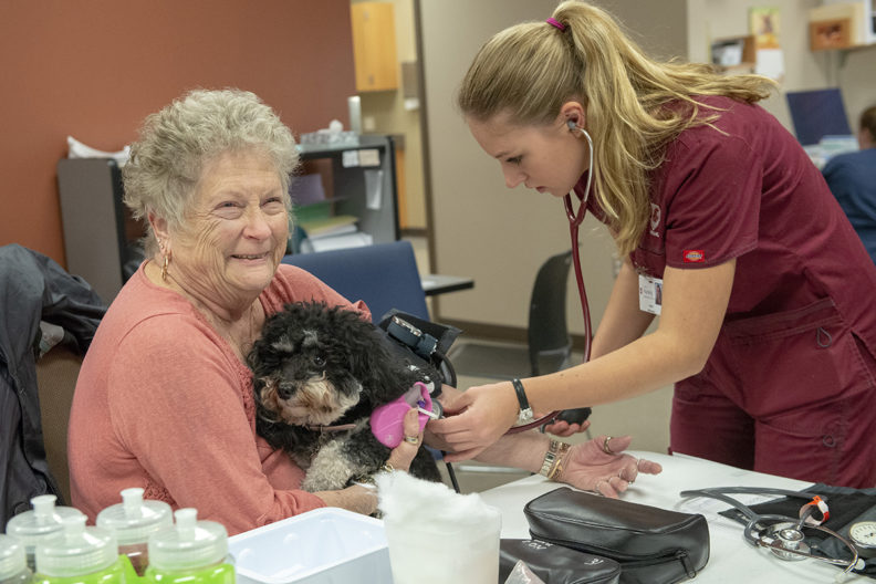 Woman holding pet dog as nursing student checks her blood pressure.