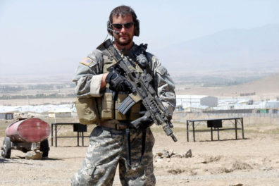 Shurer in Afghanistan in combat uniform and holding an machine gun.