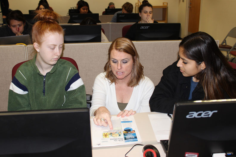 Instructor between two students with laptops discussing library project.