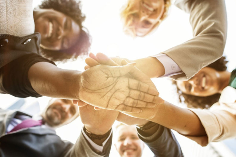 Five people of various cultures laying one hand on top of another in middle of group huddle.