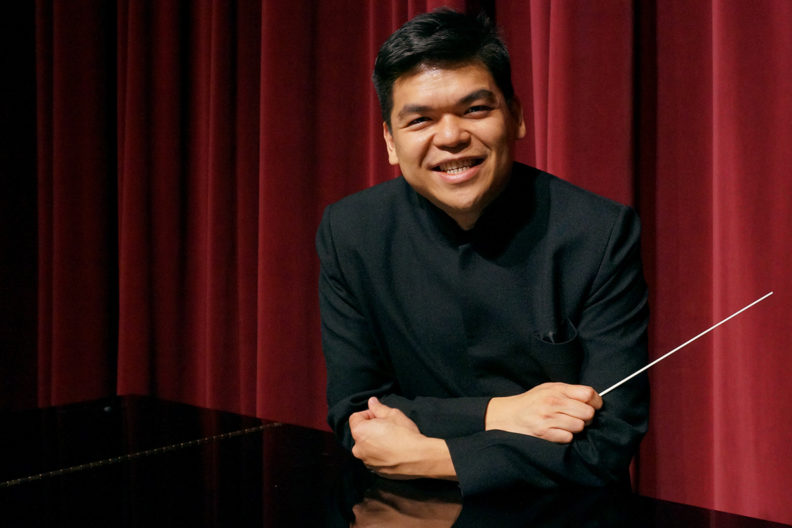 Closeup of Pham leaning against piano with baton in hand.