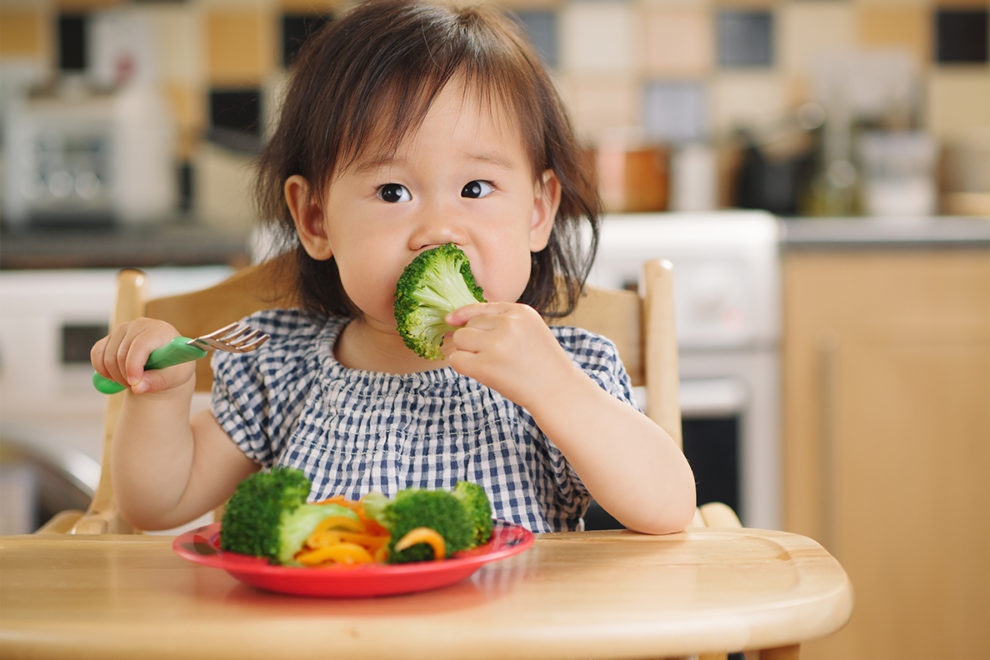 Toddler in high chair eating broccoli.