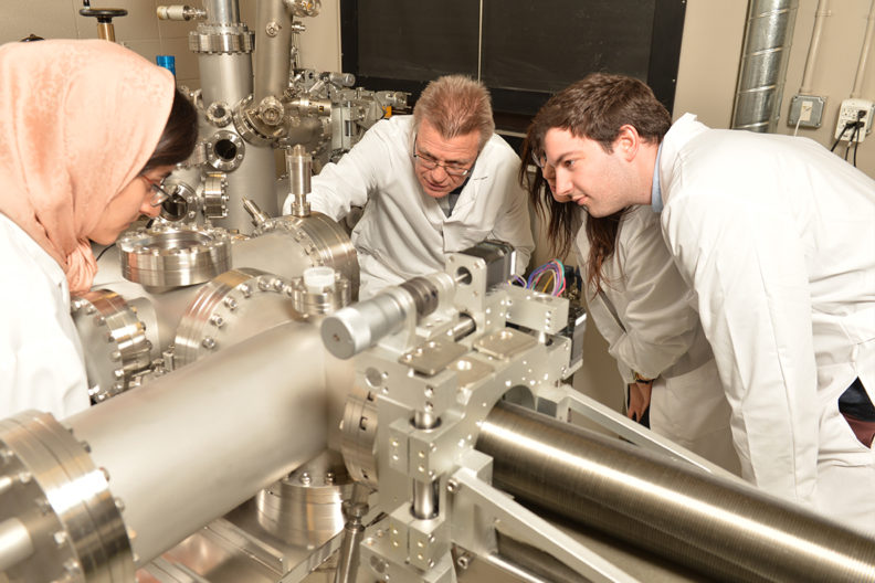 A scientist wearing a lab coat shows two other scientists how a machine is used while another scientist uses it.