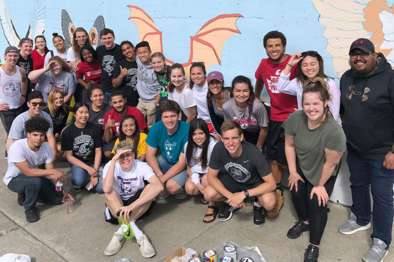 A large group of CCE student volunteers stand in front of an outdoor mural.