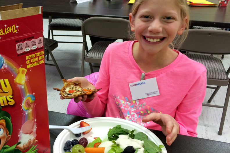 Young girl with a plate of fruit and vegetables and a box of cereal.
