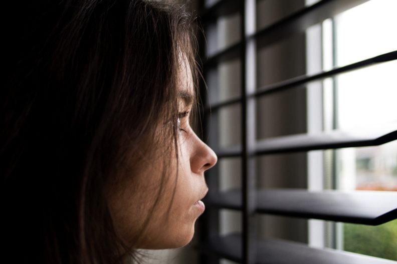 Bored girl looking through window blinds.