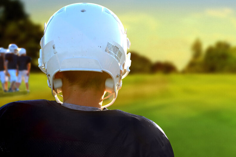 Young football player in a white helmet and blue jersey standing before an empty field.