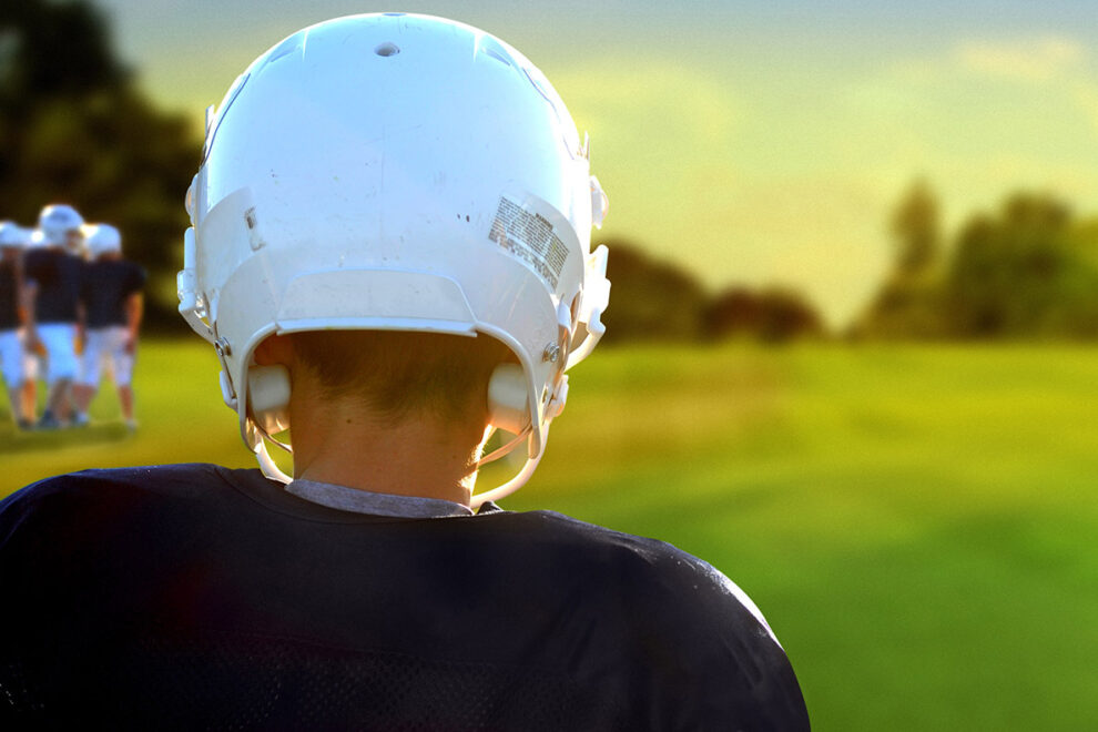 Young football player in a white helmet and blue jersey standing before an empty field.
