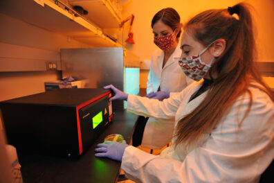 Two women in lab coats in a lab. Woman in foreground is holding a petri dish and before a boxlike irradiation machine