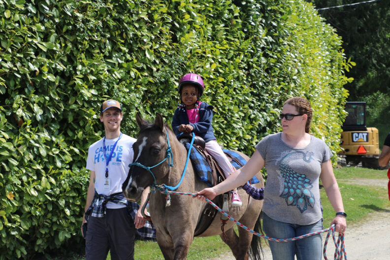 A young girl rides a horse while two camp counselors help