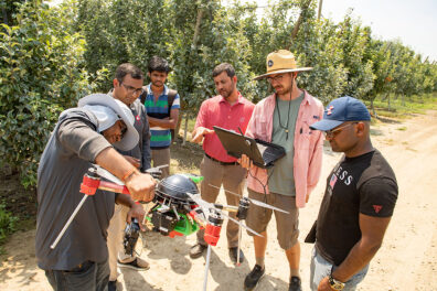 Six people standing in an orchard and inspecting a drone.